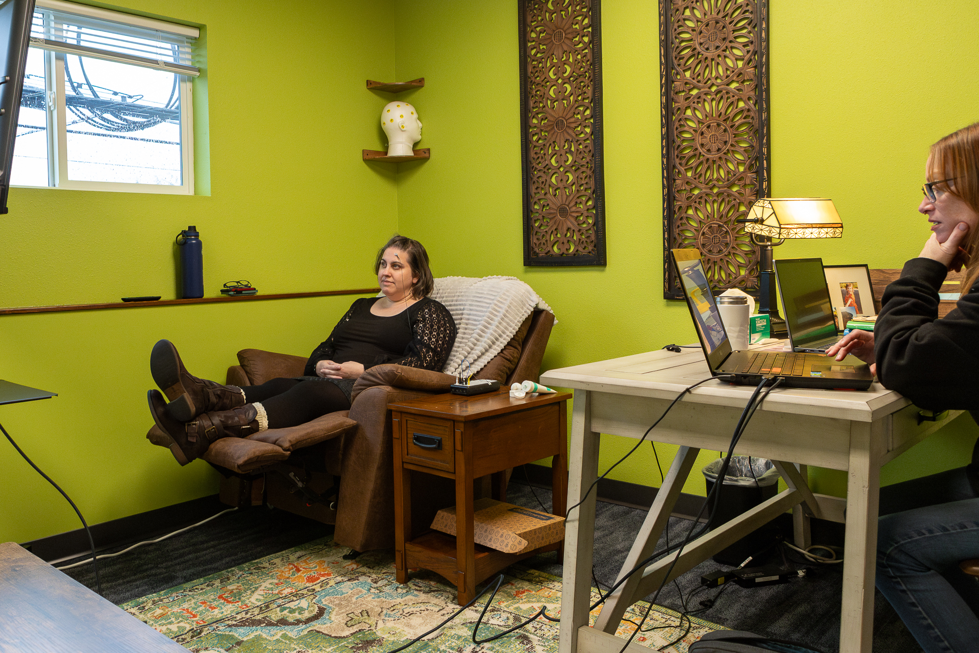 WB_Neuro_18 Client seated in a recliner with neurobiofeedback sensors attached, while a clinician monitors data on a laptop in a calm therapy room with green walls and decorative panels.